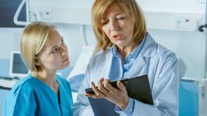 Professional Female Doctor and Young Nurse Discuss Patient's Chart. In the Background Sick Man Lying in the Bed Waiting to be Cured.